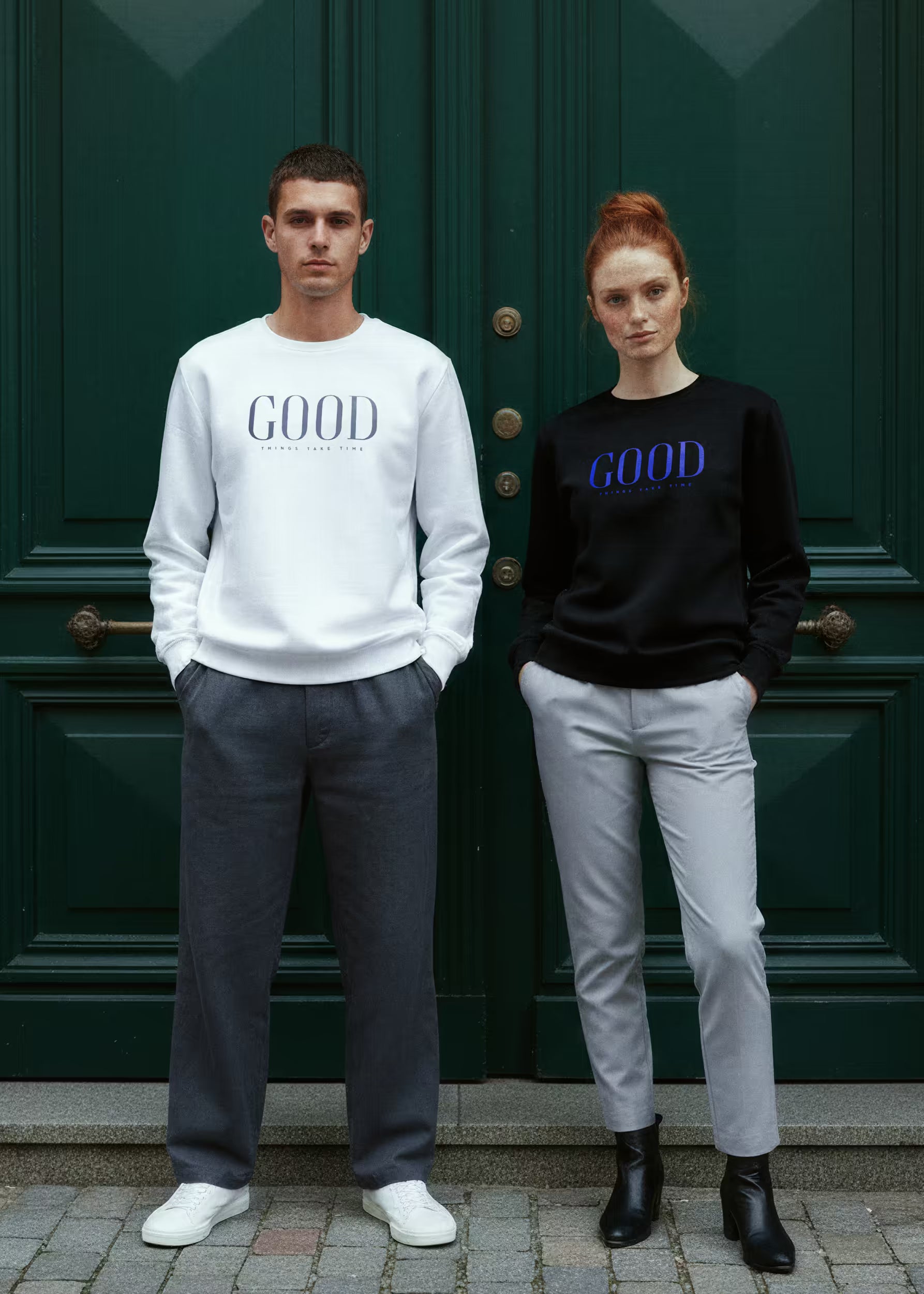 Dos modelos con sudaderas Good Things Take Time blanca y negra frente a puerta verde — Crystal Peppers