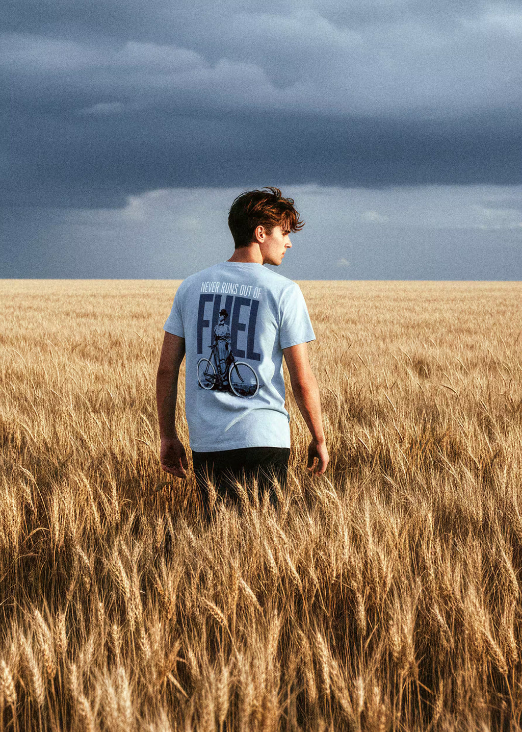 Modelo masculino con camiseta azul celeste Never Runs Out of Fuel en campo de trigo — serigrafía trasera con mujer y bicicleta, Crystal Peppers