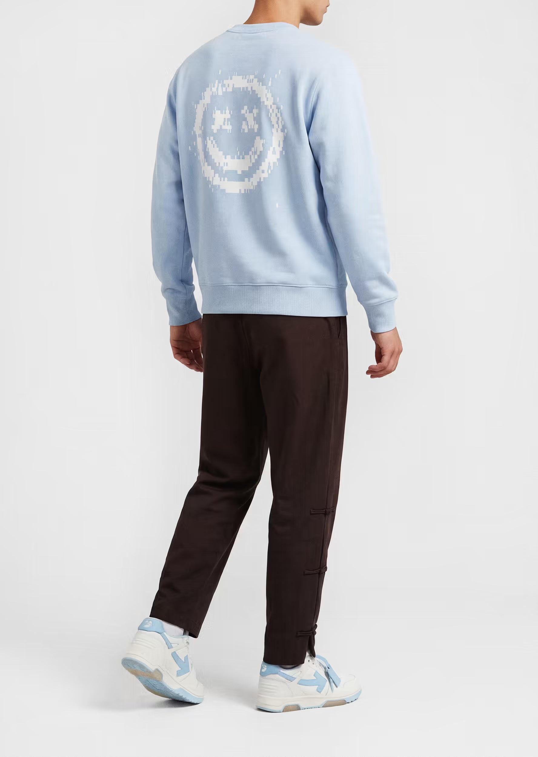 Modelo masculino con sudadera Smiley Pixel Art azul celeste y pantalón marrón — detalle de la serigrafía smiley pixelada blanca en la espalda
